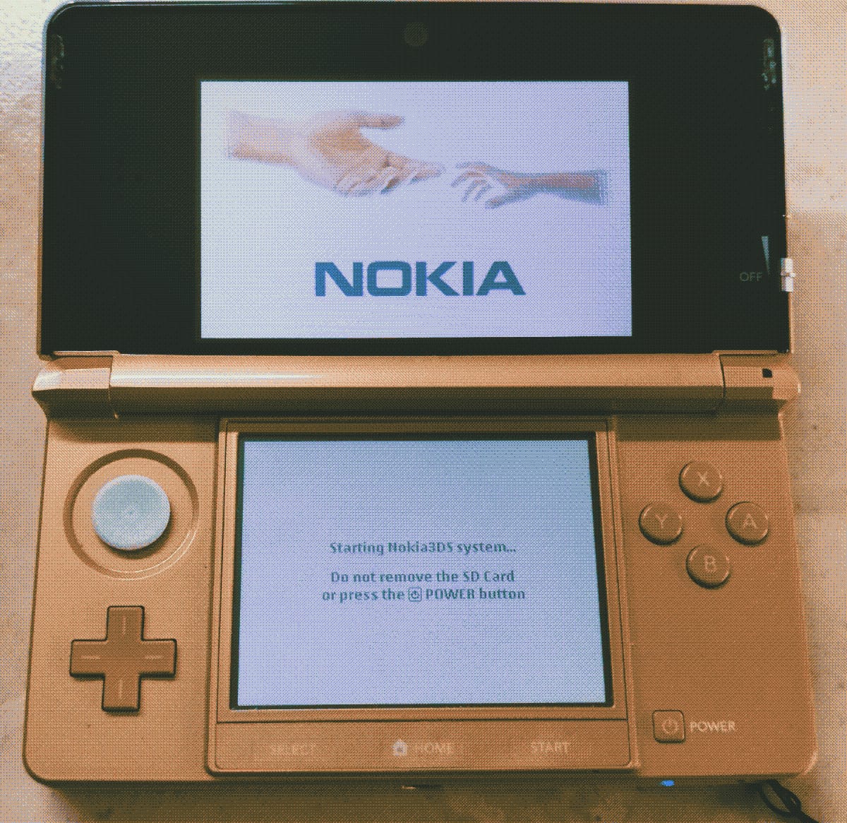 Picture of my pink 3DS, open, showing the Nokia splash screen. Top screen shows two hand reaching towards eachother with the Nokia logo underneath them. Bottom screen says: Starting Nokia3DS system... Do not remove the SD card or press the power button.